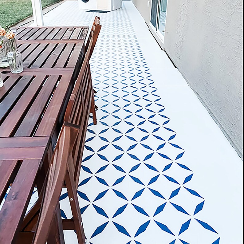 May include: Outdoor patio with a white floor featuring a blue geometric pattern. A dark brown wooden table and chairs are on the left. The floor pattern transitions from solid blue shapes to a dotted design.