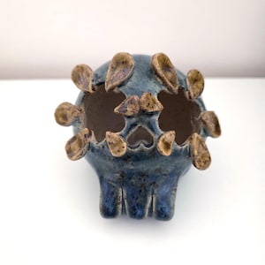 May include: A blue ceramic skull-shaped planter with a glossy, speckled finish. The top of the skull features several small, brown, petal-like protrusions arranged around the opening, and the interior is a light, unglazed beige color.
