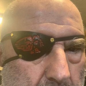 Peut inclure: Un homme portant un cache-œil en cuir noir avec un motif rouge. Le cache-œil est doté de rivets dorés.