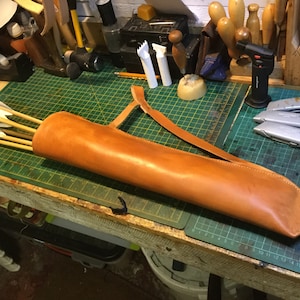 May include: A brown leather quiver with a strap, holding a set of arrows with white fletching. The quiver is lying on a green cutting mat with a ruler.