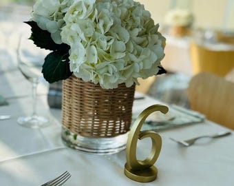May include: A table setting with a white tablecloth, featuring a centerpiece of white hydrangeas in a woven basket vase. A gold number 6 table number and a green patterned napkin with a photo are also present.