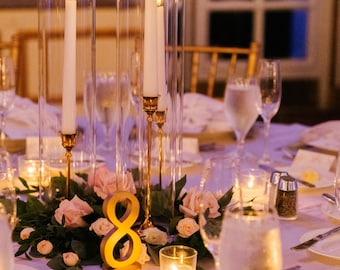May include: Elegant table setting with tall glass candle holders, lit candles, and floral arrangements. The centerpiece includes a gold number eight, surrounded by pink roses and greenery. Wine glasses and place settings complete the formal dining scene.