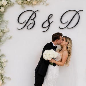 Op de afbeelding: Een huwelijksscène met een kussend stel voor een witte achtergrond versierd met bloemstukken en de letters "B & D". De bruid draagt een witte kanten jurk en de bruidegom een zwart pak. De scène speelt zich buiten af.