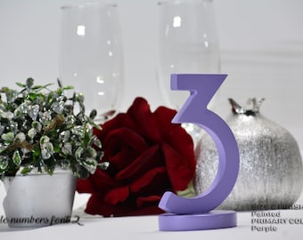 May include: A lavender painted wooden table number three, with a matching base. The number is displayed with a red rose, a silver decorative pomegranate, and a small potted plant. The text "table numbers font 2" is visible.