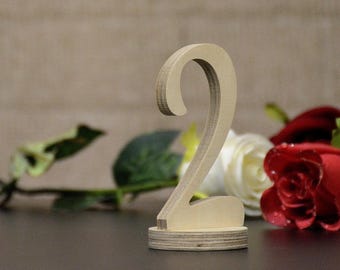 May include: A wooden number two table decoration. The number is light brown and stands on a round base. Red and white roses with green leaves are in the background. The background is a light brown color.