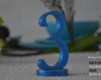 May include: A metallic blue number three wedding glitter sign. The number is painted with a metallic finish and stands on a small circular base. The sign is displayed on a table with blurred background elements.