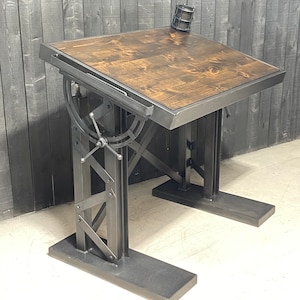 May include: A dark gray metal drafting table with a wood top. The table has a crank to adjust the angle of the top. The table is on a white floor.