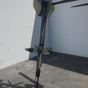 May include: A dark green and black missile-shaped sculpture with yellow accents. The sculpture has a cylindrical body with angled fins and a pointed tip. The sculpture is standing on a concrete surface.