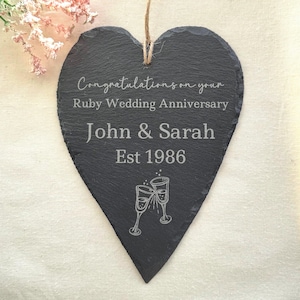 May include: A heart-shaped, dark grey slate plaque with the text "Congratulations on your Ruby Wedding Anniversary John & Sarah Est 1986" and two champagne glasses. A twine loop is attached for hanging.