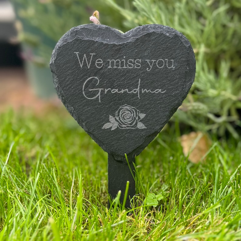 May include: A heart-shaped, black slate garden marker with the engraved text "We miss you Grandma" and a rose illustration. The marker is mounted on a black stake and placed in green grass.
