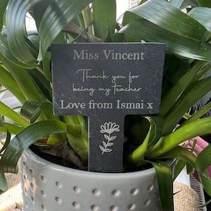 May include: A dark grey slate plant marker with the inscription "Miss Vincent, Thank you for being my teacher, Love from Ismai x" and a floral illustration. The marker is in a grey ceramic pot with a plant.