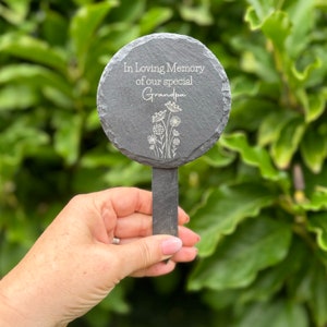 May include: A dark gray slate garden marker with the text "In Loving Memory of our special Grandpa" and a floral design. The round marker is attached to a rectangular stake.