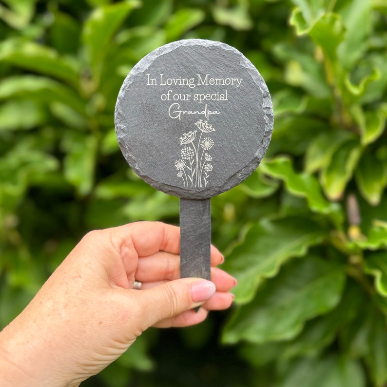 May include: A dark gray slate garden marker with the text "In Loving Memory of our special Grandpa" and a floral design. The round marker is attached to a rectangular stake.