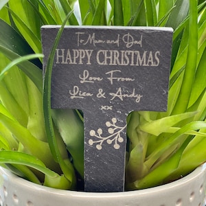 May include: A black slate plant marker with the inscription "To Mum and Dad HAPPY CHRISTMAS Love From Lisa & Andy". The marker features a floral design and is placed in a green plant, inside a cream pot.