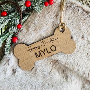 May include: A wooden dog bone Christmas ornament, engraved with "Happy Christmas MYLO". The light brown bone-shaped decoration hangs by a jute string. Red berries and pine needles are in the background.