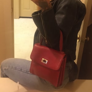 Puede incluir: Un bolso de cuero rojo con cierre de plata. El bolso se lleva sobre el hombro de una persona que lleva una chaqueta de cuero negra y pantalones vaqueros azules.