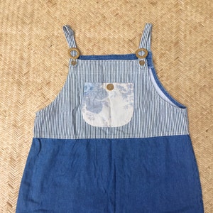 Può includere: Salopette in denim a righe blu e bianche con una tasca a stampa floreale. La salopette ha due chiusure a bottone in ottone sugli spallacci.