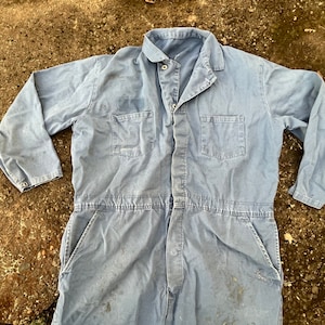 May include: A light blue work jumpsuit with long sleeves and a button-up front. The jumpsuit has two chest pockets and two large side pockets.