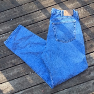 Puede incluir: Un par de pantalones vaqueros azules de mezclilla con un bolsillo trasero. Los pantalones están doblados por la mitad y colocados sobre una superficie de madera.