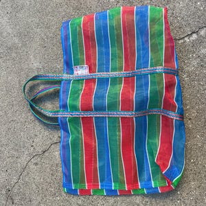 May include: A colorful tote bag with vertical stripes in red, green, and blue. The bag has a rectangular shape with a handle and a decorative trim. The bag is made of a mesh-like material.