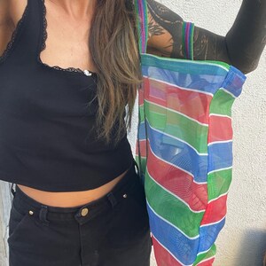 May include: A colorful mesh tote bag with horizontal stripes in red, blue, green, and white. The bag has a rainbow-striped handle and is held up against a white wall. The bag is a reusable shopping bag.