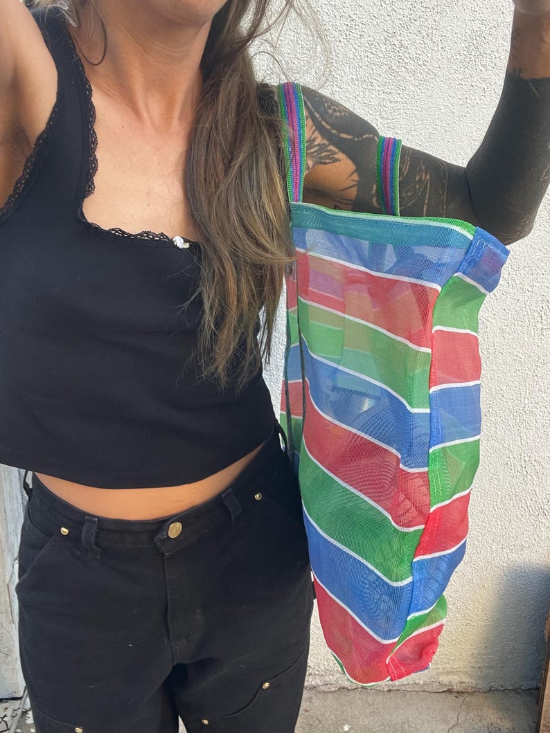 May include: A colorful mesh tote bag with horizontal stripes in red, blue, green, and white. The bag has a rainbow-striped handle and is held up against a white wall. The bag is a reusable shopping bag.
