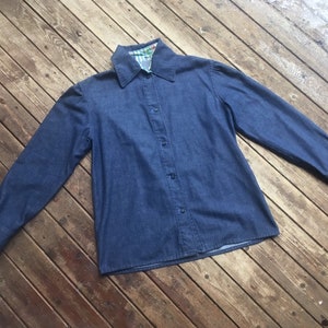 May include: A blue denim shirt with a long sleeve and a button-up front. The collar is lined with a green and white patterned fabric.