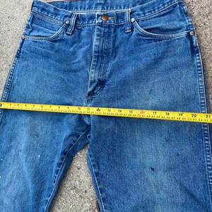 May include: Blue denim jeans with a straight leg cut. A yellow measuring tape is laid across the jeans, showing a measurement of approximately 58 cm. The jeans have a button closure and classic five-pocket design.