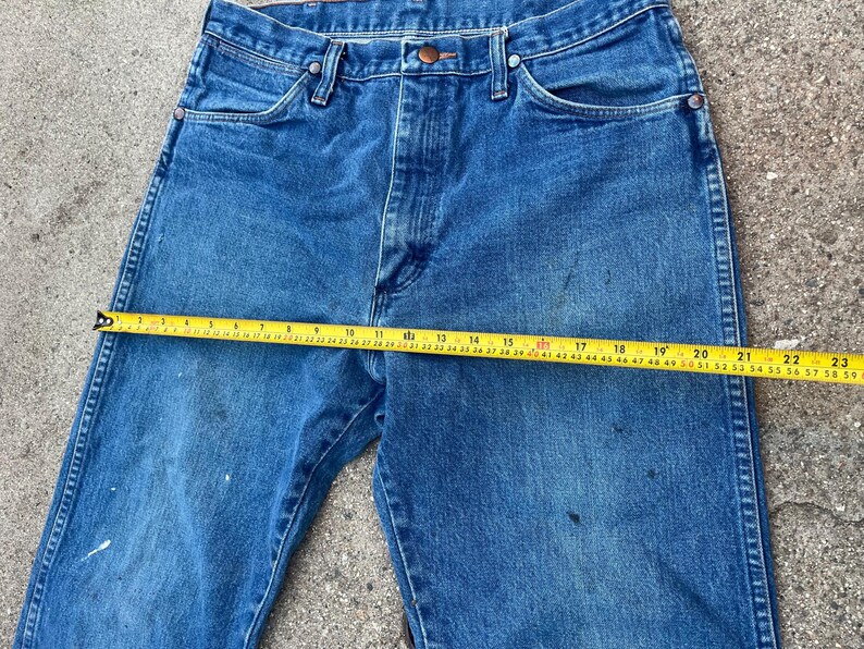 May include: Blue denim jeans with a straight leg cut. A yellow measuring tape is laid across the jeans, showing a measurement of approximately 58 cm. The jeans have a button closure and classic five-pocket design.