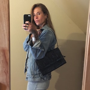 May include: A woman wearing a denim jacket and jeans is holding a black purse with a long strap. The purse has a textured design.