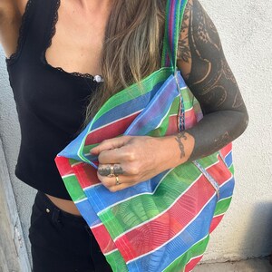 May include: A colorful, striped tote bag with a rainbow pattern of red, green, blue, and purple. The bag has a matching striped strap and is made of a mesh-like material. The bag is held up by a person.