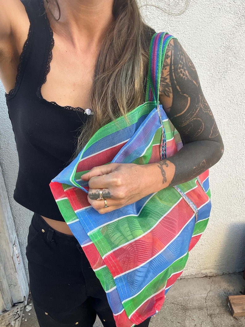 May include: A colorful, striped tote bag with a rainbow pattern of red, green, blue, and purple. The bag has a matching striped strap and is made of a mesh-like material. The bag is held up by a person.