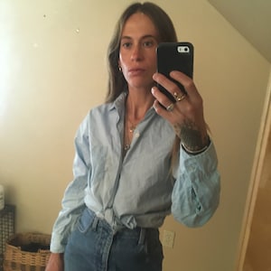 May include: A woman wearing a light blue button-down shirt tied at the waist and a denim mini skirt. She is holding a black smartphone and wearing gold rings.