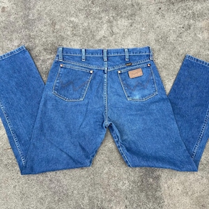 Puede incluir: Un par de pantalones vaqueros azules de mezclilla con un parche de cuero marr&oacute;n en el bolsillo trasero que dice "Wrangler".