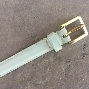 May include: A white leather belt with a gold buckle. The belt is stitched with a white thread.