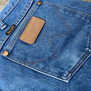 Puede incluir: Primer plano del bolsillo trasero de un par de pantalones vaqueros de mezclilla azul. El bolsillo tiene un parche de cuero marr&oacute;n con la palabra "Wrangler" cosida en blanco.