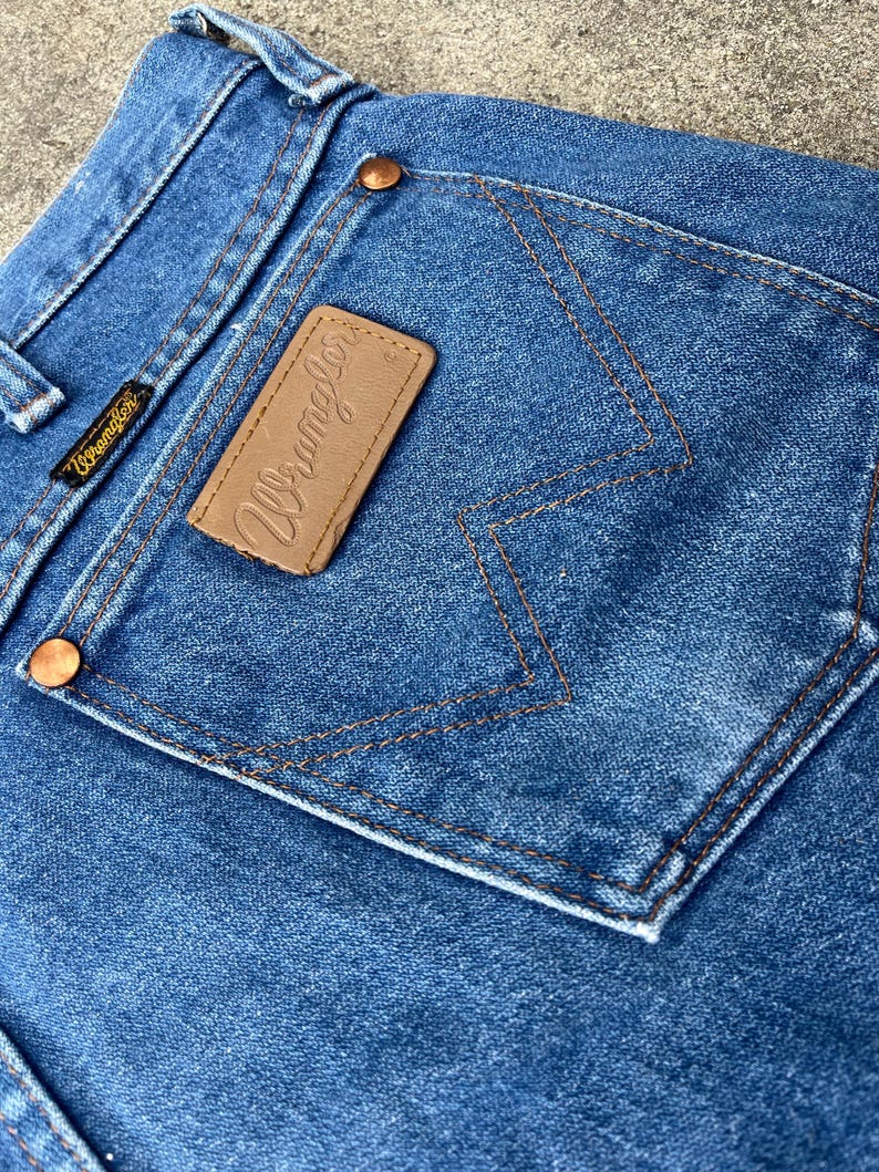 Puede incluir: Primer plano del bolsillo trasero de un par de pantalones vaqueros de mezclilla azul. El bolsillo tiene un parche de cuero marr&oacute;n con la palabra "Wrangler" cosida en blanco.