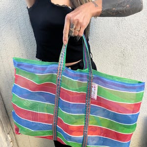 May include: A large, colorful striped tote bag with red, blue, green, and white horizontal stripes. The bag has braided green handles and a small tag. The bag is made of a sheer material.