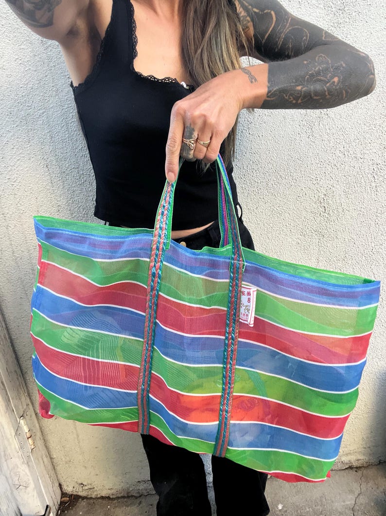 May include: A large, colorful striped tote bag with red, blue, green, and white horizontal stripes. The bag has braided green handles and a small tag. The bag is made of a sheer material.