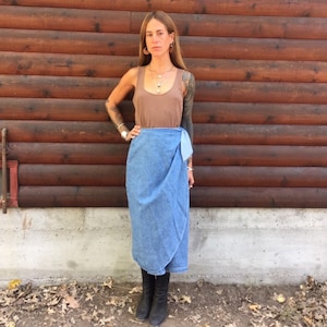 May include: A woman wearing a brown tank top and a blue denim wrap skirt. She is standing in front of a wooden wall.