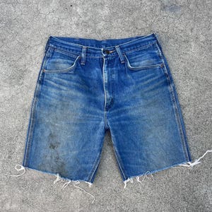 May include: Blue denim shorts with a faded wash and raw, frayed edges. The shorts have classic five-pocket styling and a button and zipper closure. The shorts appear to be vintage or pre-worn, with some discoloration.