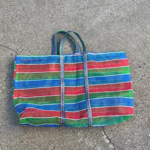 May include: A large, rectangular tote bag with a vibrant striped pattern in red, blue, green, and white. The bag has two sturdy handles and is made of a mesh-like material. The bag is lying on a concrete surface.
