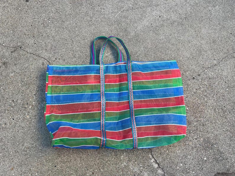 May include: A large, rectangular tote bag with a vibrant striped pattern in red, blue, green, and white. The bag has two sturdy handles and is made of a mesh-like material. The bag is lying on a concrete surface.