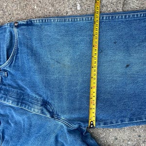 May include: Close-up of blue denim jeans with a measuring tape showing a 91 cm waist. The jeans are a medium wash with visible wear and tear. The tape measure is yellow with black markings.