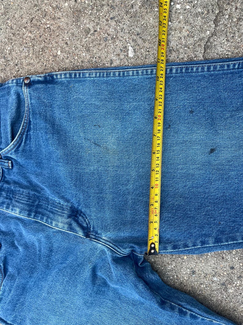 May include: Close-up of blue denim jeans with a measuring tape showing a 91 cm waist. The jeans are a medium wash with visible wear and tear. The tape measure is yellow with black markings.