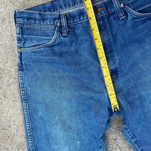 May include: Blue denim jeans with a yellow measuring tape showing a measurement of approximately 35.5 cm. The jeans have a classic five-pocket design with metal rivets and a button closure. The denim has some wear and tear.