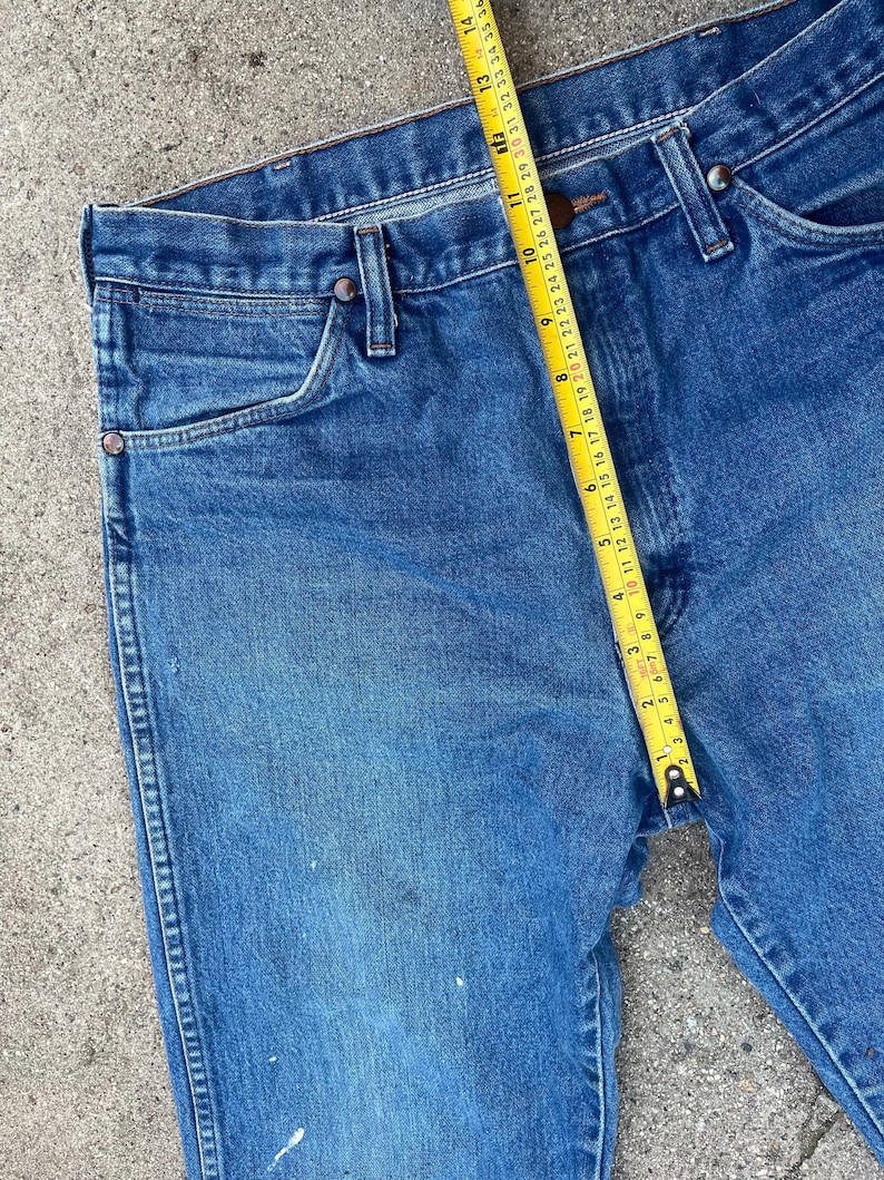 May include: Blue denim jeans with a yellow measuring tape showing a measurement of approximately 35.5 cm. The jeans have a classic five-pocket design with metal rivets and a button closure. The denim has some wear and tear.