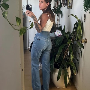 May include: A woman wearing a white vest top and light blue jeans with a tool pocket on the left leg. She is standing in front of a mirror and looking at her reflection.