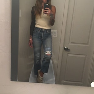 Könnte beinhalten: Eine Frau trägt ein weißes Tanktop und eine blaue Jeans mit Destroyed-Details. Sie steht vor einem Spiegel und hält ein Handy.