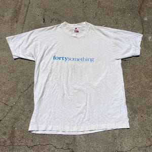 May include: White t-shirt with the text "fortysomething" in blue across the chest. The shirt has short sleeves and a crew neck. The fabric appears slightly wrinkled.